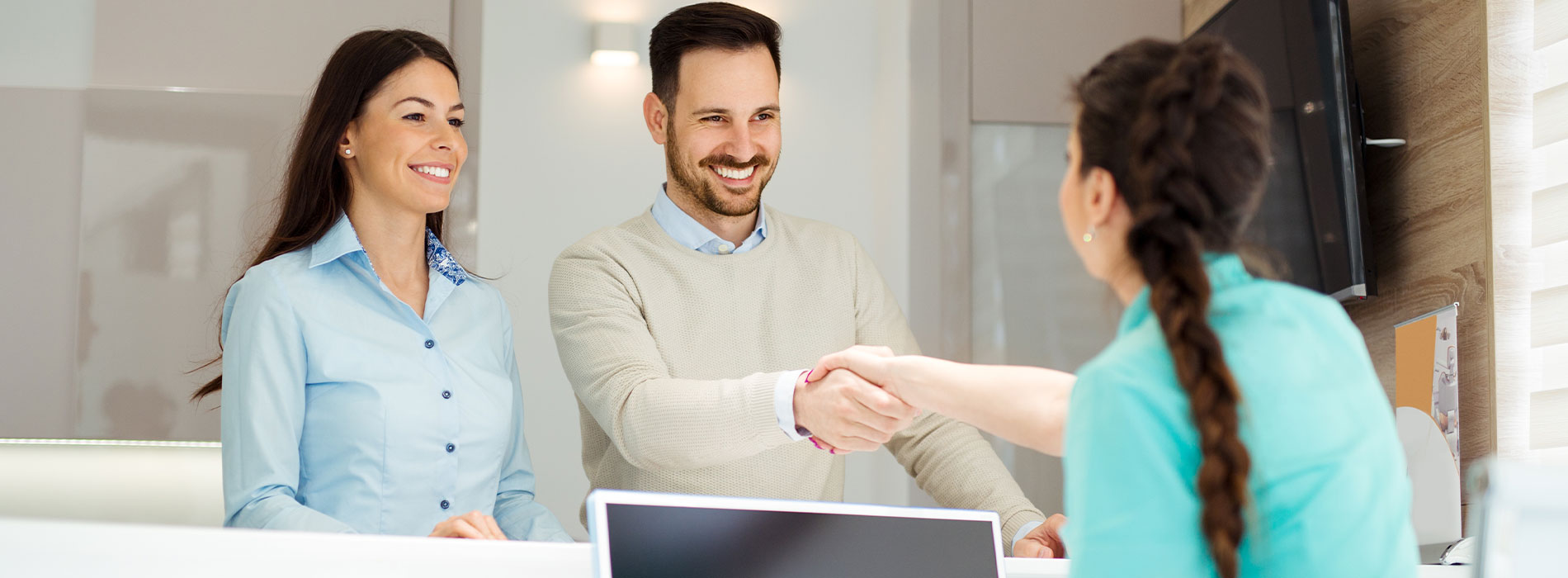 Two people shaking hands in a professional setting, with another person observing behind them.