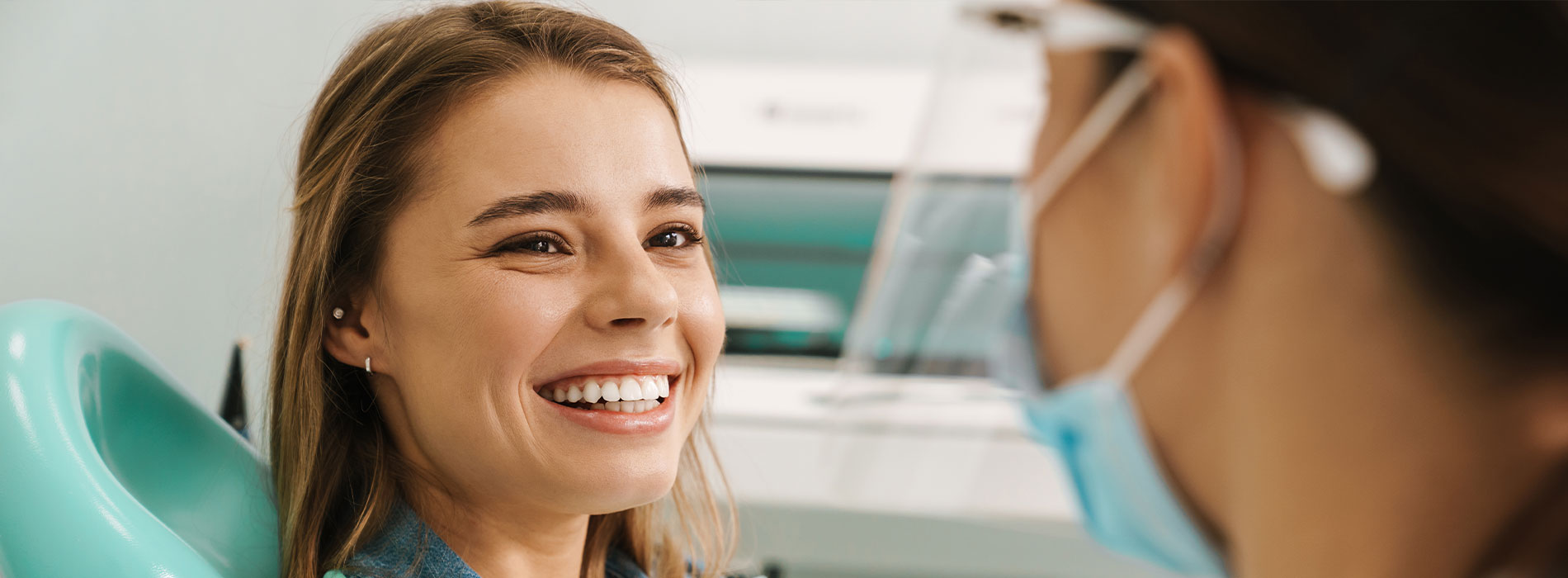 The image shows a young woman with her mouth open, sitting in a dental chair, smiling at another person who appears to be a dental professional, with both individuals wearing medical masks and surrounded by dental equipment.