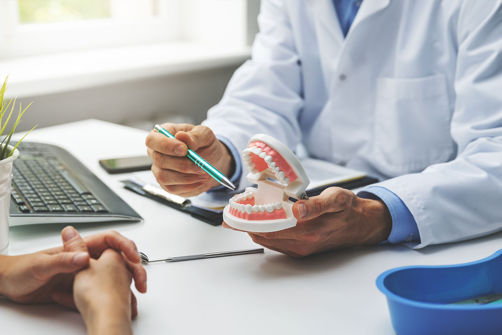 A dental professional holding a model set of teeth, examining them with a magnifying glass while seated at a desk.