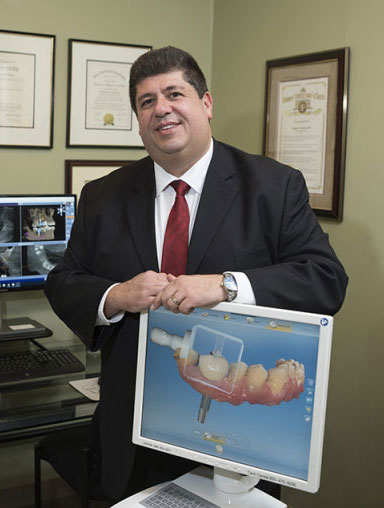 The image shows a man standing behind a desk with a computer monitor displaying a 3D model of a human head, holding a model of a tooth implant, smiling slightly at the camera.