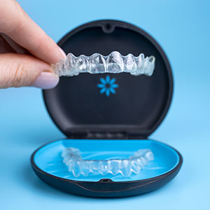 The image shows a person's hand holding a clear plastic tray containing a set of dental braces with visible teeth impressions, placed inside an open blue case with a white circular logo on the lid, which appears to be a brand name.