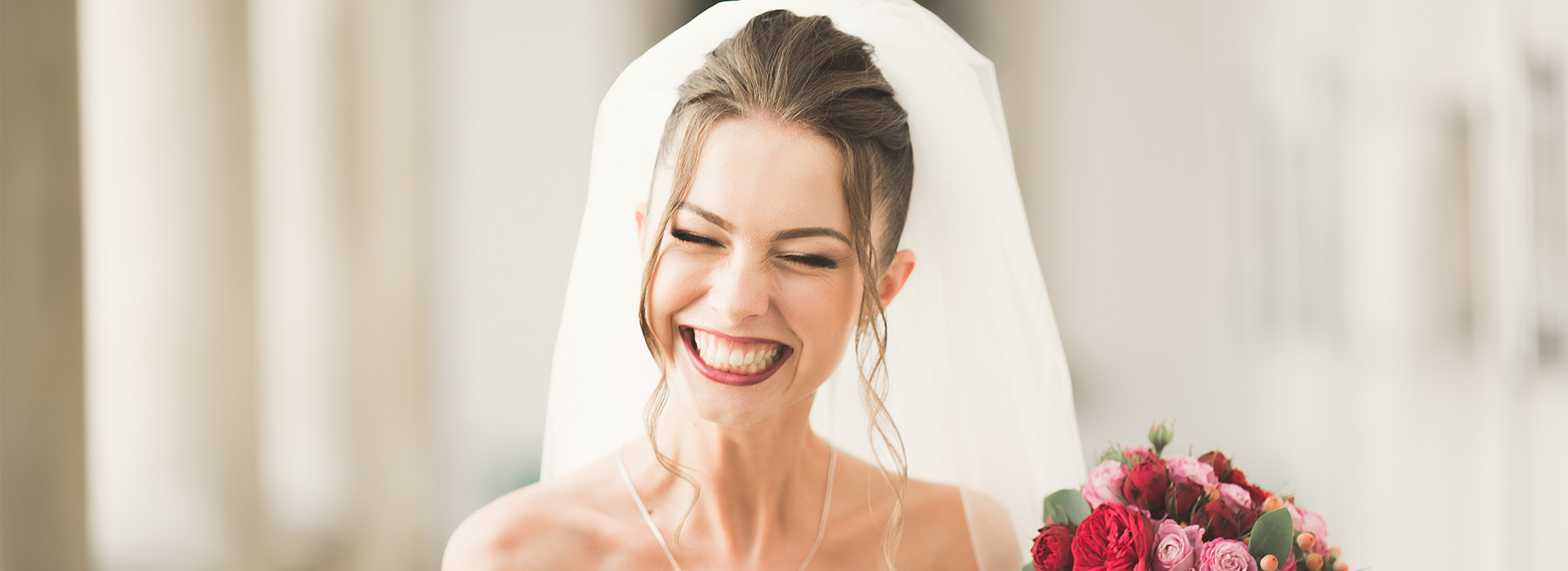 A bride with joyous expression, wearing a white veil and holding flowers, smiling at the camera.