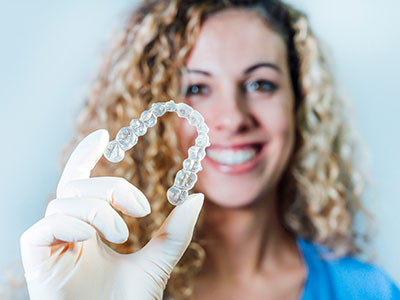 A smiling woman holding up a clear dental retainer with her gloved hand.
