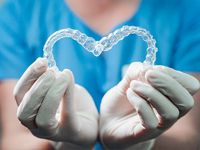 A person wearing medical gloves holds a transparent heart-shaped object with intricate details, symbolizing dental braces, against a blue background.