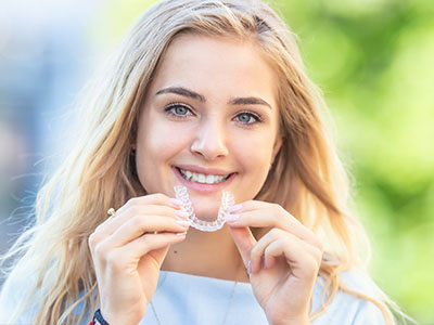 A young woman with blonde hair smiling at the camera while holding a clear dental retainer in her hand.
