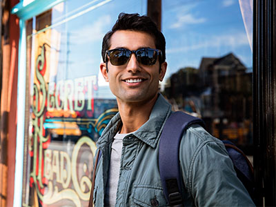 A man wearing sunglasses and a backpack stands confidently against a storefront window with a neon sign.
