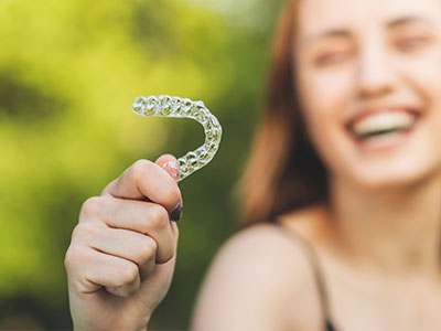 A young woman holding up an orthodontic retainer with a smile on her face, set against a blurred outdoor background.