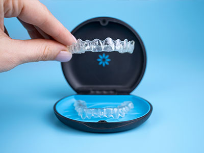 An image showcasing a transparent plastic dental retainer with a blue lid, being held up by a human hand against a light blue background.