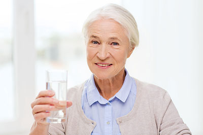 The image features an elderly woman holding a glass of water, smiling slightly, with her arm extended towards the camera.