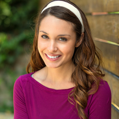 A young woman with long brown hair, wearing a purple top, smiling at the camera against a wooden fence background.