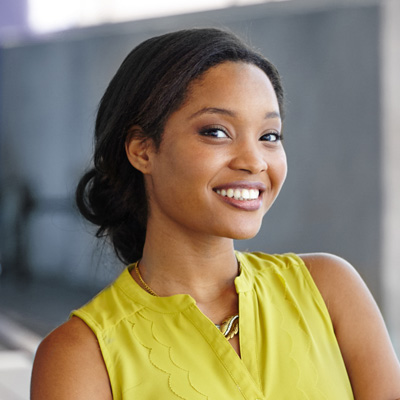The image shows a woman with a smile, posing against a background wall, wearing a yellow top and a necklace.