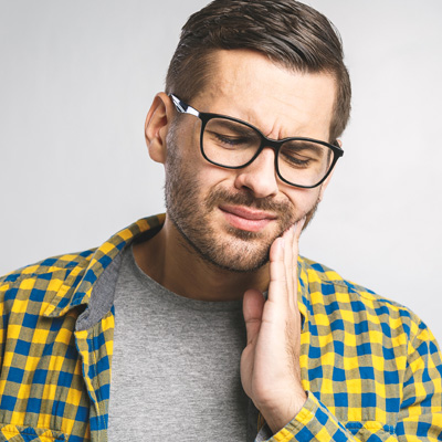 A man with glasses and a beard wearing a yellow plaid shirt, looking upwards with his hand on his chin, showing signs of discomfort or pain.
