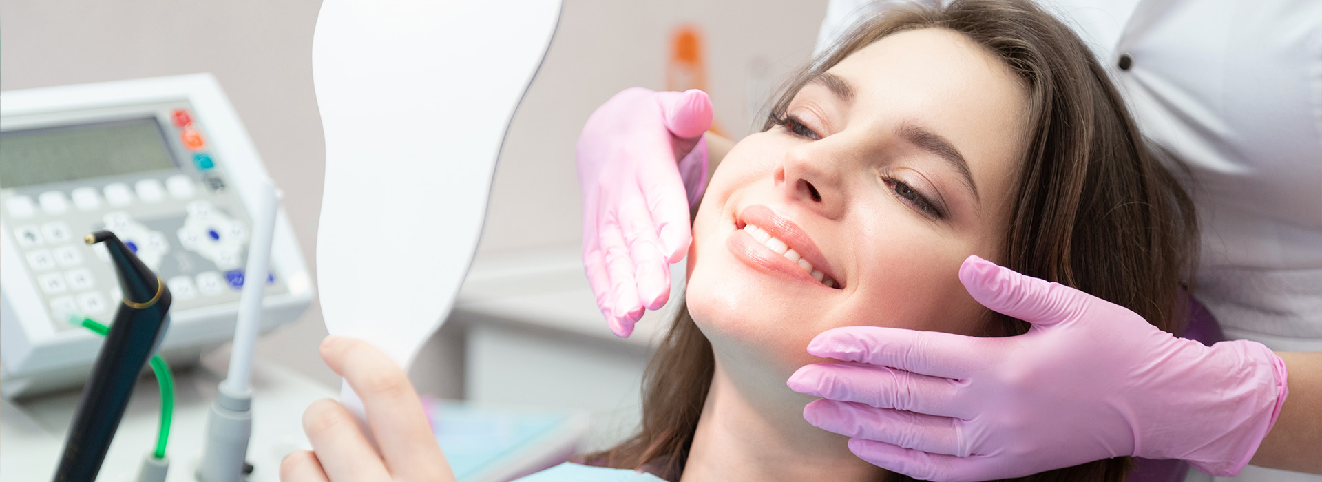 The image shows a woman receiving dental care, with a dentist performing an examination using a mirror, while she sits in the dental chair with her eyes closed, wearing a pink face mask and a dental headrest.