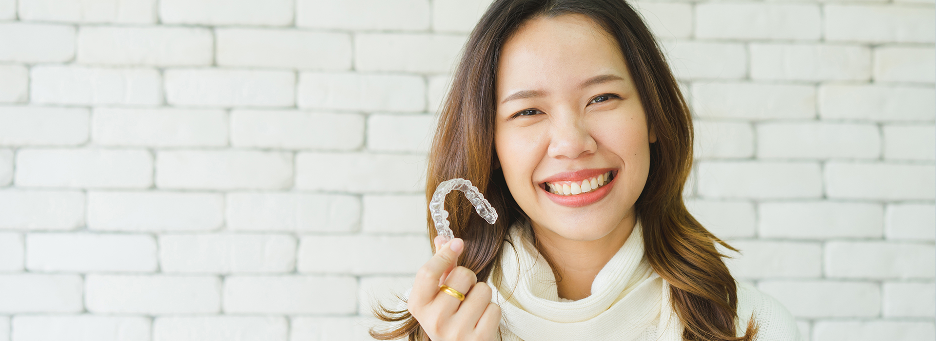 A woman with a smile is holding a ring, standing against a brick wall background.