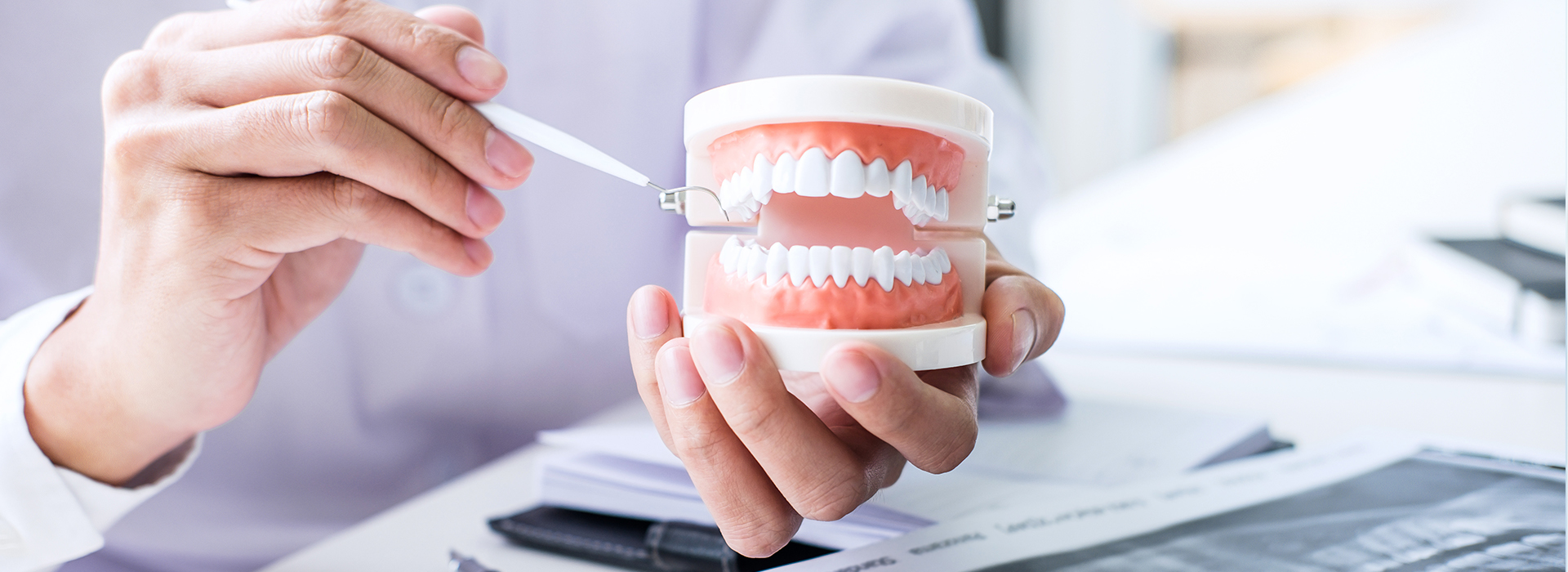 A person's hand holding a toothbrush with white bristles next to a cup of toothpaste, against a blurred background of an office setting.