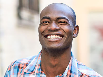 The image shows a smiling man with short hair, wearing a plaid shirt, standing outdoors against a backdrop that includes a building facade and a clear sky.
