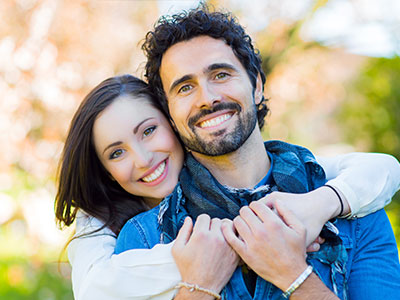 A man and woman are hugging each other with smiles, set against an outdoor background.