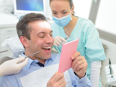 A man sitting in a dental chair with a pink cardboard sign on his chest, smiling broadly at the camera while a female dentist looks on with a surgical mask and gloves.