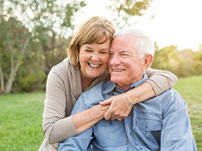 The image shows an elderly couple embracing each other outdoors during daylight, with both individuals smiling at the camera.