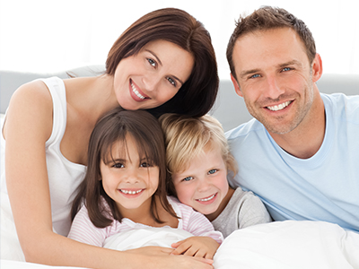The image depicts a family of five people lying together on a bed with smiles on their faces.