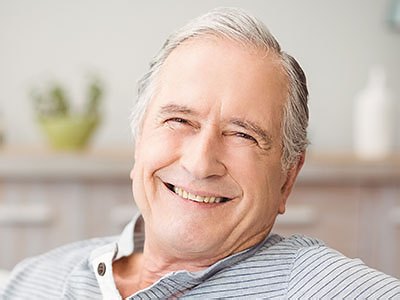 The image shows an older man with gray hair smiling at the camera while sitting comfortably in a chair.
