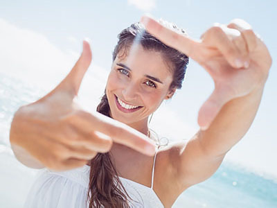 A young woman with long hair smiles while taking a selfie with her hand held up against a sunny beach backdrop.